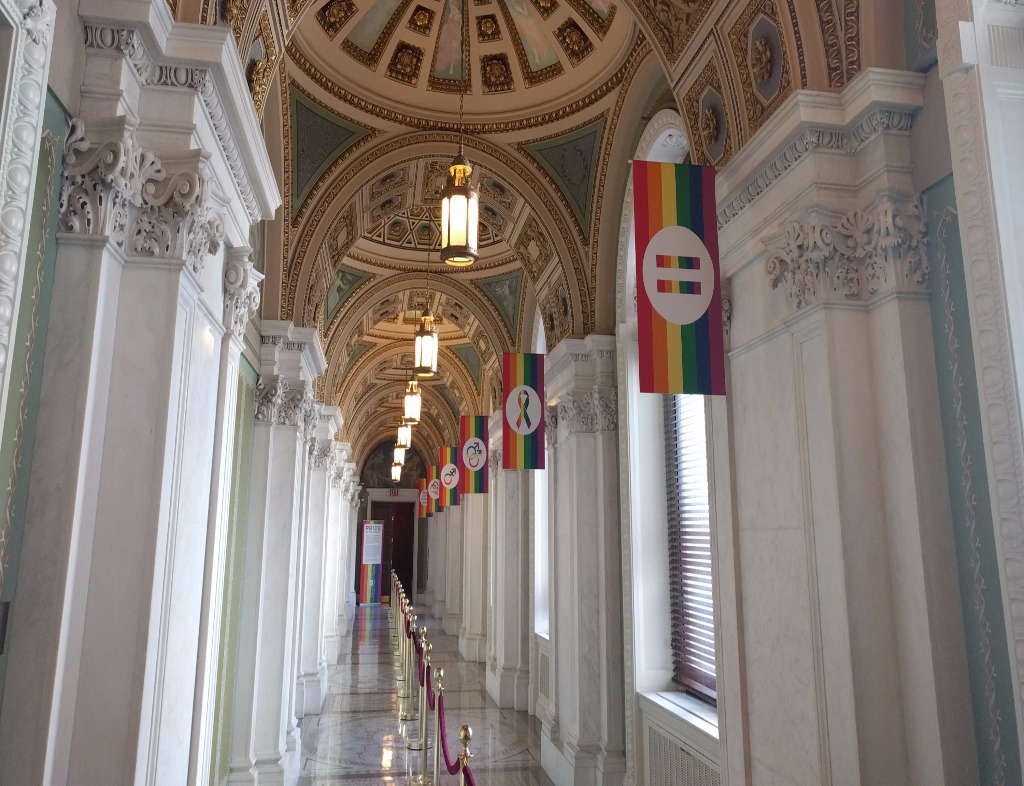 The view down a hallway in the Jefferson Building of the Library of Congress. Marble floor, marble walls and pillars, and a series of dome ceilings. There are windows along the right side, each with a rainbow flag and different LGBTQ-related symbols. A velvet rope divides the hallway lengthwise.