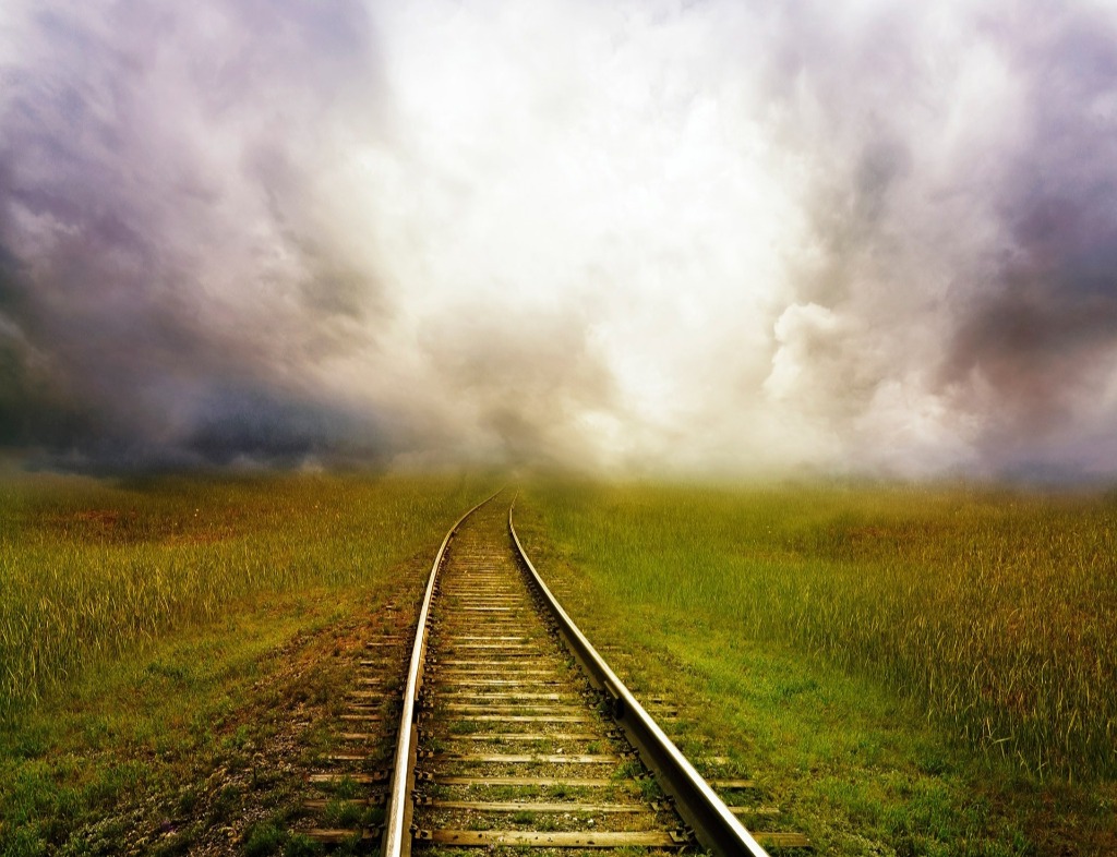 Train tracks at the center of the photo heading into very thick, gray clouds, although the sun shines from behind them. The tracks are on top of grass.