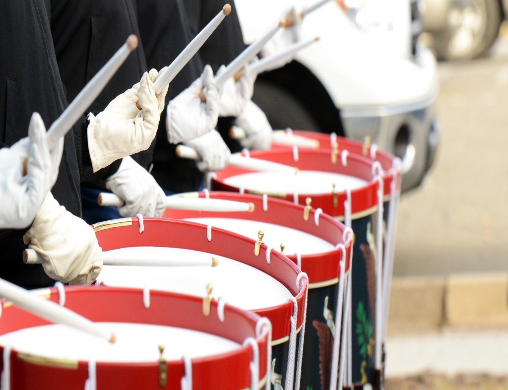 A line of five field drums being played be musicians with white gloves, white drumsticks, and black or dark blue outifts. The drumheads are white, the drums are red at the rim and have a black or dark blue body.